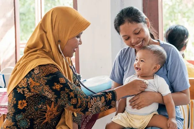 Kader kesehatan mendengarkan seorang ibu yang menggendong bayinya di posyandu desa