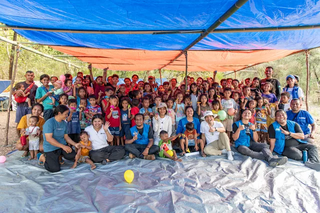 Dozens of residents and children pose with a volunteer team under a tarpaulin shelter at an emergency response site