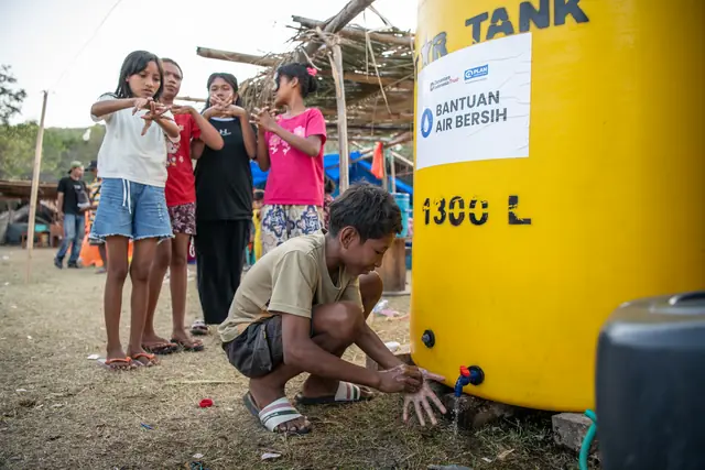 Children wash their hands at an aid water tank alongside a laughing volunteer