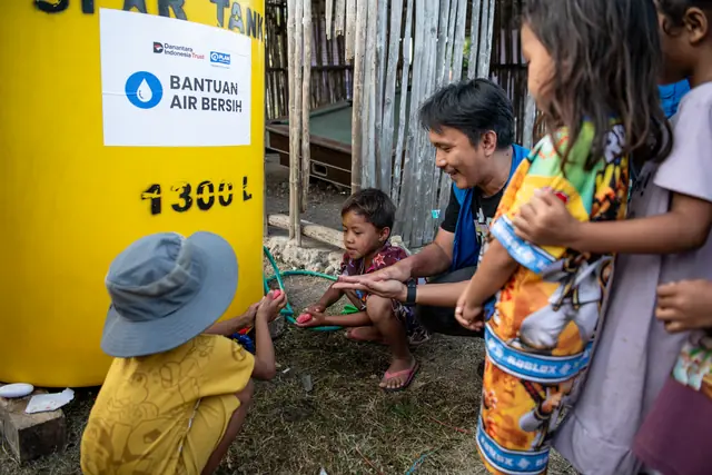 A volunteer helps a child in a bucket hat wash their hands at a Danantara aid water tank